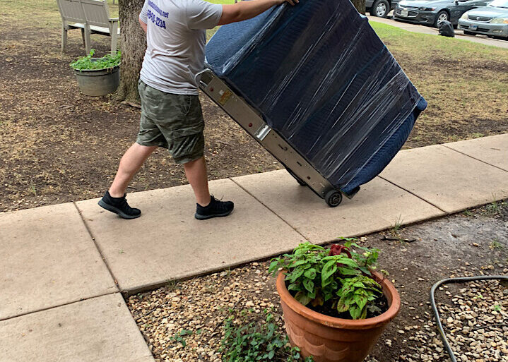 mover-pushing-wrapped-furniture-on-dolly A professional mover pushing a large piece of furniture wrapped in blue plastic on a dolly along an outdoor sidewalk
