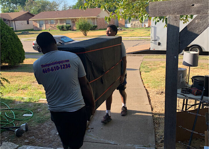 movers-carrying-furniture-outdoors Two movers carrying a large piece of furniture wrapped in black blankets along a sidewalk