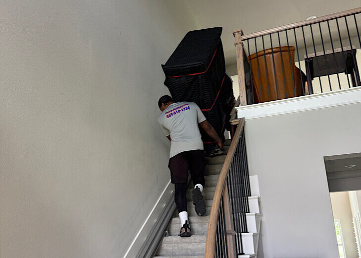 movers-carrying-protected-furniture-upstairs Two professional movers carefully carrying a large piece of furniture wrapped in black protective blankets up a curved residential staircase