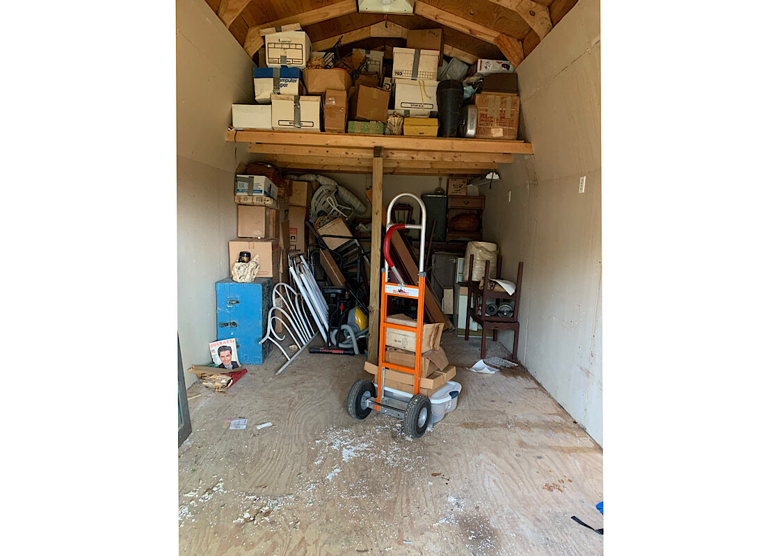 Dallas Shed Cleanout Service Interior of a cluttered wooden storage shed in Dallas filled with boxes and furniture, featuring an orange moving dolly ready for junk removal